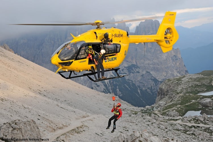 Un hombre cuelga de un cable desde un helicóptero amarillo, con las montañas al fondo.