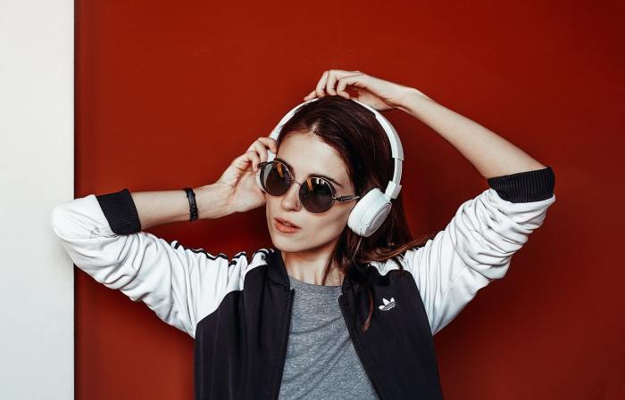 Una chica con gafas oscuras escucha música con auriculares blancos frente a un fondo rojo.