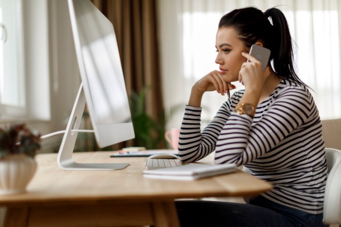 Una mujer está sentada frente a su computadora, al mismo tiempo que está haciendo una llamada telefónica.