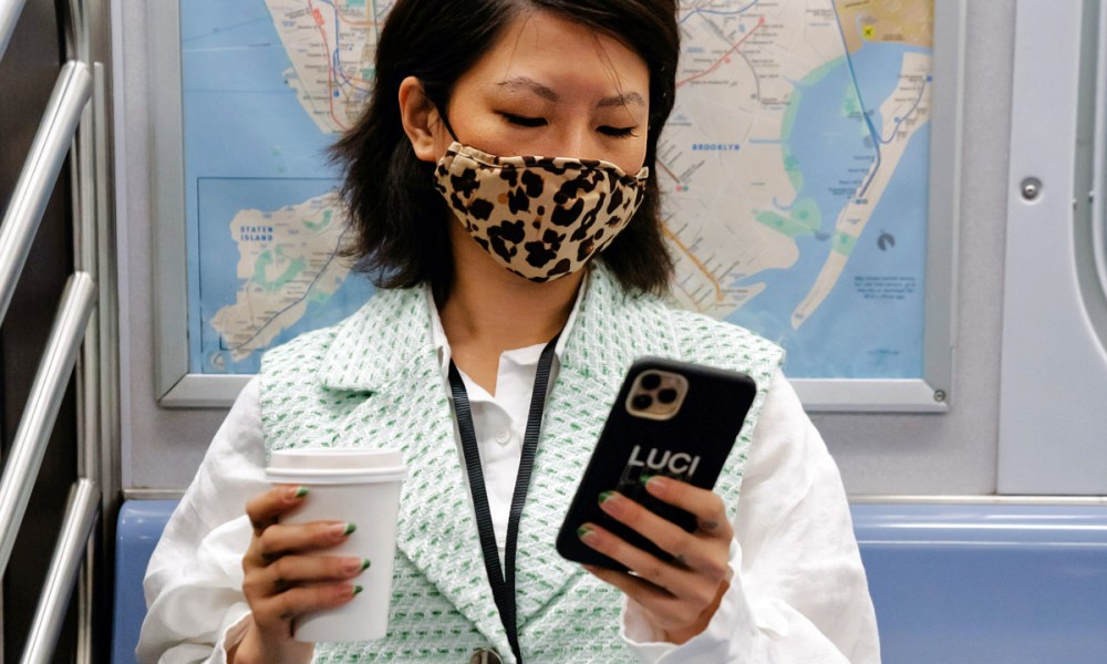 Una mujer en el metro y con mascarilla observa la pantalla de su iPhone mientras sostiene un café con la otra mano.