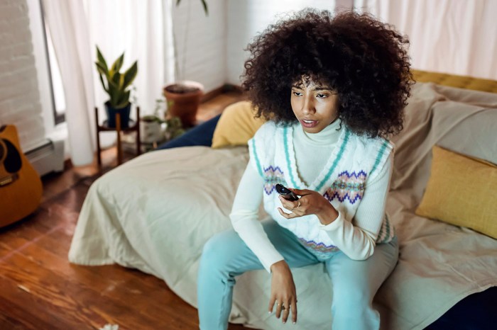Una chica de gran cabello rizado sentada en su cama con el control remoto del televisor en la mano y expresión aburrida.