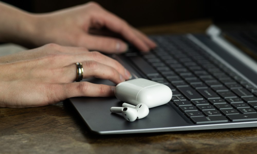 Mujer tecleando en una MacBook con unos AirPods sobre esta – Cómo conectar los AirPods a la MacBook sin perderte en el intento